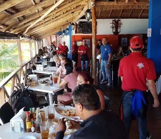 A bustling seaside restaurant with patrons dining at tables under a wooden thatched roof. People are engaged in conversation and enjoying their meals, many discussing their upcoming spring break Cancun dates. A waiter in a red shirt is serving drinks. Tropical decor and ocean views add to the vibrant atmosphere.