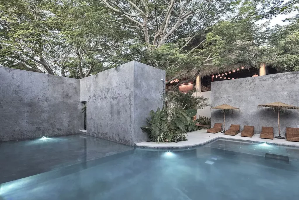 Modern outdoor pool area with concrete walls, surrounded by lush trees. Several lounge chairs with straw umbrellas are set beside the pool, reminiscent of an idyllic Bacalar oasis in Mexico. Greenery accents the scene, making it an inviting retreat for 2024.