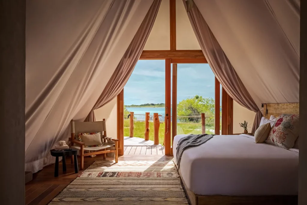 Glamping tent interior with a double bed, armchair, and deck overlooking a grassy area and tranquil body of water under the blue sky of Bacalar, Mexico.
